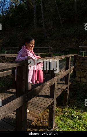 Une femme dans une chemise rose se tient sur un pont en bois. Elle sourit et regarde au-dessus d'un plan d'eau. Le pont est entouré d'arbres, et le soleil Banque D'Images