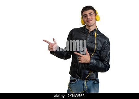 Jeune homme avec des écouteurs jaunes et une veste en cuir noir pointant avec les index vers la gauche sur fond blanc Banque D'Images