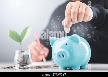 Un homme d'affaires dépose une pièce de monnaie dans une tirelire bleue, coin jar avec un petit semis poussant au centre. Il écrit des chiffres dans un cahier, symbolisant Banque D'Images
