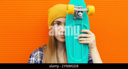 Portrait de jeune fille élégante avec skateboard en chapeau jaune, adolescente moderne posant sur fond orange Banque D'Images