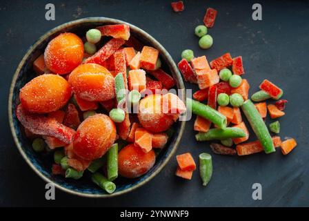 Un mélange vibrant de légumes surgelés dans un bol, mettant en valeur leurs couleurs vives sur un fond bleu profond. Banque D'Images