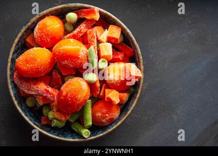 Pois, tomates, carottes et poivrons fraîchement congelés disposés dans un bol, soulignant leurs textures sur une surface bleu foncé. Banque D'Images