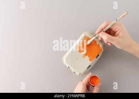 Peindre un carton d'oeufs orange avec un pinceau pour un projet artisanal de bricolage sur un fond gris. Fabrication des mains en cours. Recyclage créatif et enfants a Banque D'Images