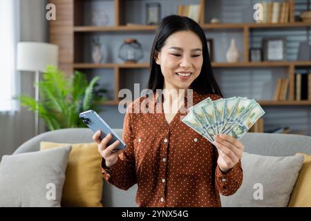 Femme asiatique dans le cadre de la maison tenant de l'argent et smartphone, souriant. Affiche le concept de la réussite financière, des gains en ligne, du bonheur et de l'utilisation de la technologie avec le téléphone. Banque D'Images