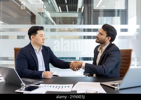 Hommes d'affaires serrant la main dans le bureau moderne. L'image illustre le partenariat, la confiance et la réussite de l'affaire. Les ordinateurs portables, les graphiques, les documents sur le bureau mettent en évidence l'environnement professionnel. Concept de travail d'équipe, coopération Banque D'Images