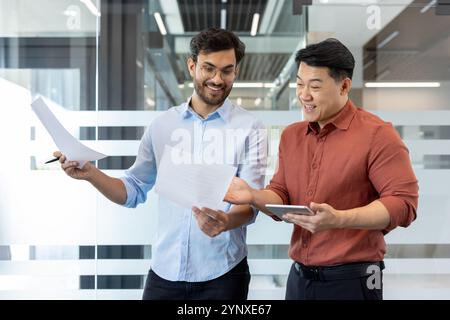 Deux collègues collaborent sur des plans de projet tout en examinant des documents et en utilisant une tablette. Accent mis sur le travail d'équipe, la communication et l'environnement de bureau productif avec des médias numériques et imprimés. Banque D'Images