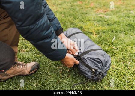 Mains de l'homme déballant un sac de tente gris sur l'herbe verte illuminé de lumière naturelle dans la chaîne de montagnes des Andes Banque D'Images