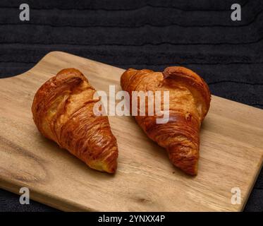 Deux croissants fraîchement cuits reposent sur une planche de bois, mettant en valeur leurs couches feuilletées et leurs croûtes brunes dorées. Le réglage confortable de la cuisine ajoute de la chaleur à t Banque D'Images