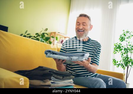 Homme senior appréciant le week-end à la maison, plier les vêtements sur un canapé jaune confortable, entouré d'une lumière naturelle rafraîchissante Banque D'Images