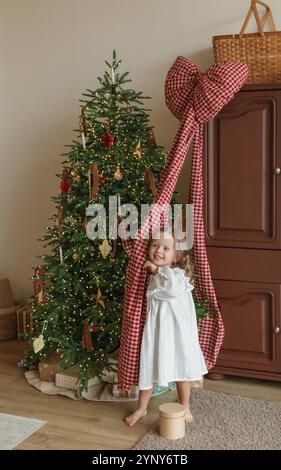 Fille souriante dans une chemise de nuit debout près d'un arbre de Noël jouant avec une décoration d'arc géant sur un placard Banque D'Images