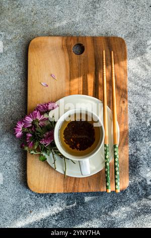Vue aérienne d'une tasse de thé sur une planche à découper en bois avec des fleurs d'aster violettes et des baguettes Banque D'Images