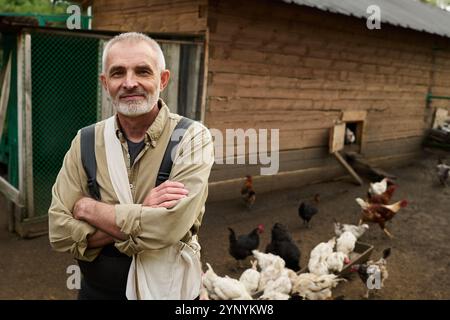 Agriculteur masculin senior en vêtements de travail regardant la caméra contre une longue coopérative en bois et un groupe de poulets picotant de l'avoine de la mangeoire Banque D'Images