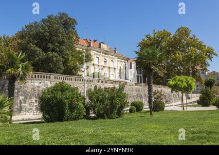 Parc paysager avec pelouse d'herbe verte et vieux bâtiment architectural jaune avec tuiles de toit en terre cuite abritant les bureaux du comté de Zadar à la fin s. Banque D'Images