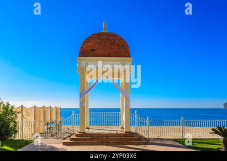 Un petit pavillon avec un auvent blanc se trouve sur une plage à côté de l'océan. Le pavillon est entouré d'une clôture et d'un banc. Le ciel est clair et bleu, Banque D'Images