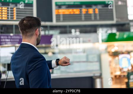 l'homme d'affaires copyspace, vu de derrière, vérifie sa montre-bracelet tout en regardant les panneaux d'information de la gare pour trouver l'heure de départ de son train, p Banque D'Images