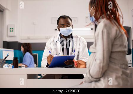 Femme noire enceinte parle avec le médecin au bureau portant tous les deux des masques faciaux pour la protection. Infirmière féminine travaille sur ordinateur en arrière-plan en manipulant des documents de santé tandis que le médecin masculin s'occupe du patient. Banque D'Images