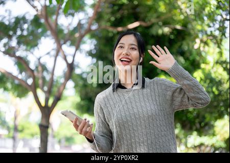 Une femme asiatique joyeuse dans des vêtements chauds vagues pour saluer ses amis tout en marchant le long du trottoir dans un parc verdoyant par une journée lumineuse, profitant de la fraîcheur Banque D'Images