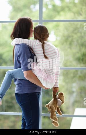 Mère, fille et vue de la fenêtre pour la curiosité à la maison, transporter l'enfant et se lier ensemble le week-end. Maman, fille et retour de famille pour la sécurité dans Banque D'Images