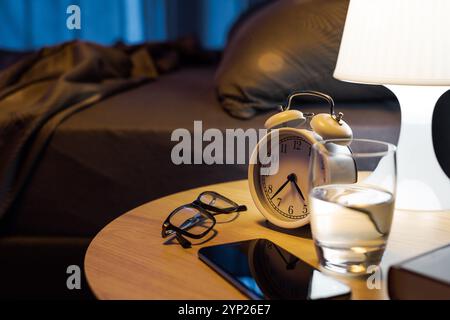 Objets personnels et lampe allumée sur la table de chevet dans la chambre, lit en arrière-plan Banque D'Images