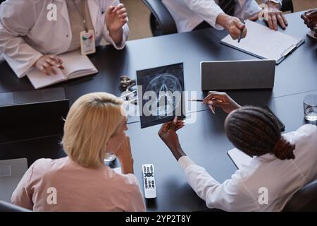 Prise de vue à angle élevé d'une femme afro-américaine tenant une image tomodensitométrique du crâne du patient discutant des résultats de l'examen radiographique avec un collègue, tout en étant assis à TAB Banque D'Images
