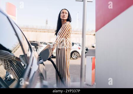 Vue latérale de la femme mature chargeant la voiture avec la prise électrique à la station Banque D'Images
