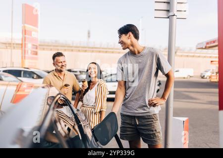 Heureux jeune homme chargeant la voiture avec la prise électrique tout en parlant avec les parents à la station Banque D'Images