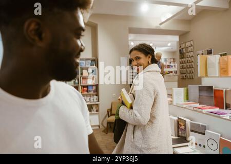 Femme souriante jetant un coup d'œil à l'homme tout en magasinant des livres à la boutique Banque D'Images