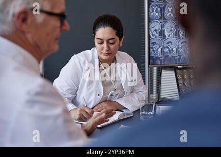 Plan moyen d'une chirurgienne du moyen-Orient réfléchie écrivant des notes sur papier planifiant le traitement des patients tout en étant assise à la table de réunion avec son collègue Banque D'Images