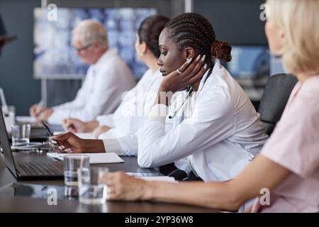 Vue de côté de clinicienne afro-américaine écoutant la présentation des collègues garder des registres de réunion de travail tout en étant assis à table dans le cabinet médical, espace de copie Banque D'Images