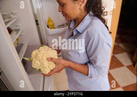 Une femme sort un chou-fleur frais du réfrigérateur, symbolisant une vie saine et une nutrition. Le cadre de la cuisine évoque un sentiment de cuisinier à la maison Banque D'Images
