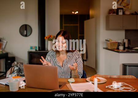 Femme freelance souriante parlant sur un appel vidéo via un ordinateur portable à la maison Banque D'Images