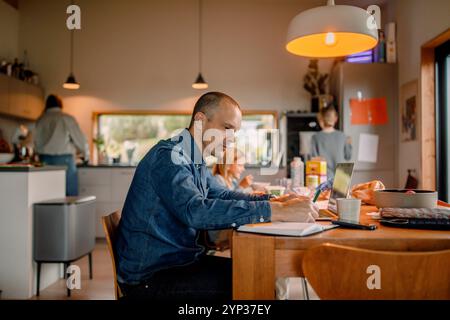Vue latérale de l'homme freelance écrivant dans le livre tout en étant assis près de la table à manger à la maison Banque D'Images