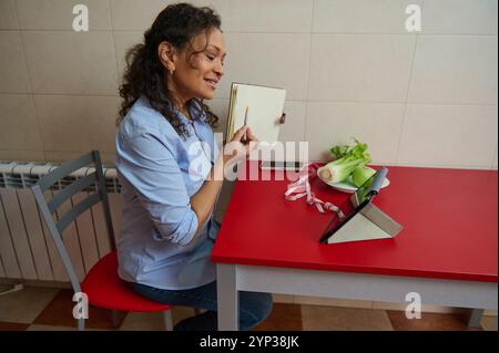 Une femme s'assoit à une table de cuisine participant à un cours de cuisine en ligne à l'aide de sa tablette. Elle tient un cahier, un stylo et sourit tout en se concentrant sur la scre Banque D'Images