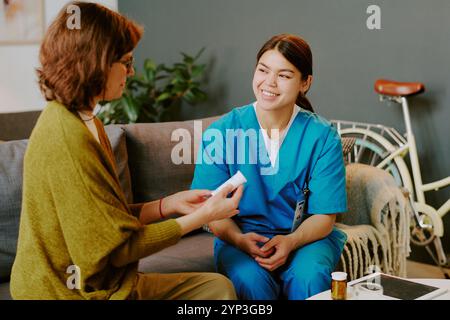 Infirmière souriante portant des gommages bleus tenant la documentation médicale tout en étant assis sur le canapé pendant la consultation du patient dans le cadre de la maison, l'environnement intérieur comprend le vélo et la plante luxuriante Banque D'Images