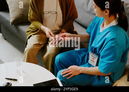 Infirmière portant des gommages bleus aidant la femme âgée avec des médicaments tout en étant assise sur le canapé dans le cadre confortable du salon avec table basse au premier plan Banque D'Images