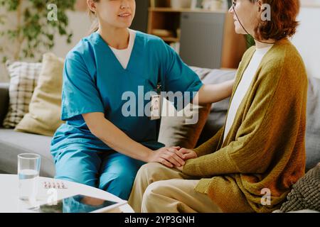 Infirmière portant des gommages bleus aidant la femme âgée dans un cadre confortable de salon, fournissant soutien et soins tout en étant assis sur le canapé Banque D'Images