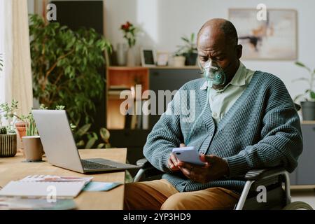 Homme senior portant un masque à oxygène tout en utilisant un smartphone et assis dans le bureau à domicile Banque D'Images