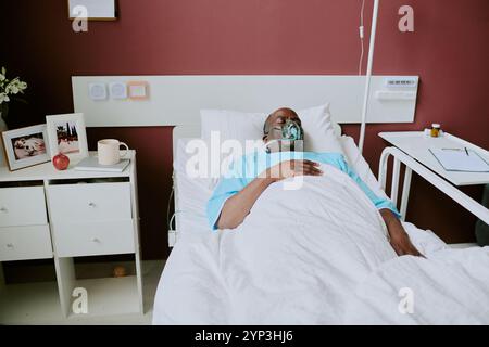 Personne âgée reposant dans un lit d'hôpital avec assistance respiratoire entourée d'équipement médical et d'articles personnels ajoutant une touche personnelle à l'environnement clinique Banque D'Images