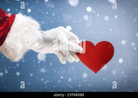 Concept d'amour de Noël avec la main du Père Noël tenant une découpe de coeur sur fond enneigé. Banque D'Images