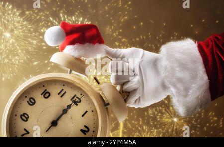 Main du Père Noël dans un costume et un gant tenant une horloge qui montre 12 heures décorée avec un chapeau de Père Noël et des feux d'artifice dorés en arrière-plan. Banque D'Images