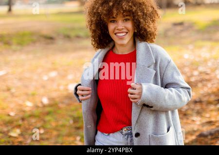 La journée d'automne lumineuse captive lorsqu'une jeune femme se promène dans un joli parc, vêtue d'un manteau gris confortable et d'un pull rouge, incarnant la chaleur et la joie au milieu Banque D'Images