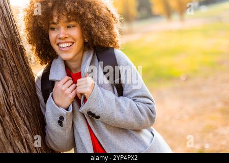 Jeune femme souriante avec les cheveux bouclés se tient contre un arbre, portant une tenue confortable. Elle embrasse la beauté de l'automne dans un parc serein, rempli de colo Banque D'Images