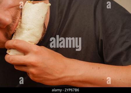 Gros plan d'un homme barbu tenant et mangeant un rouleau de viande avec des légumes frais. Capture l'anticipation de la première morsure Banque D'Images