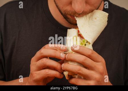 Gros plan d'un homme barbu tenant et mangeant un rouleau de viande avec des légumes frais. Capture l'anticipation de la première morsure Banque D'Images