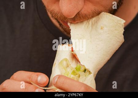 Gros plan d'un homme barbu tenant et mangeant un rouleau de viande avec des légumes frais. Capture l'anticipation de la première morsure Banque D'Images