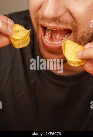 La main d'un homme tient des chips près de sa bouche lorsqu'il mange. Parfait pour les collations et les marques alimentaires. Un homme mange avec les deux mains en même temps Banque D'Images