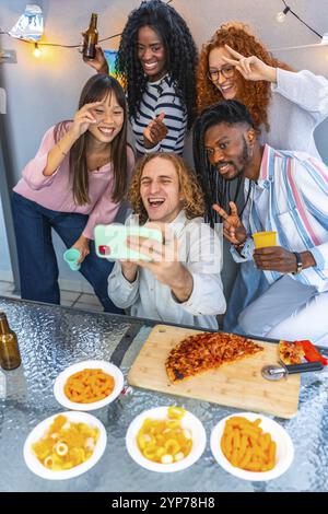 Photo verticale d'un groupe d'amis heureux multi-ethniques faisant des gestes en prenant selfie dans une fête sur le toit Banque D'Images