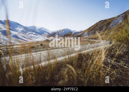 Route sinueuse mène à travers un paysage montagneux herbeux et enneigé sous un ciel bleu, Lindis Pass, Nouvelle-Zélande, Océanie Banque D'Images