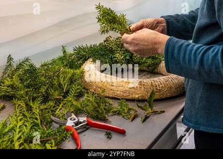 Une personne dispose soigneusement de la verdure fraîche et des cônes sur une base de couronne dans un atelier bien éclairé, se préparant pour la saison des fêtes avec créativité et f Banque D'Images