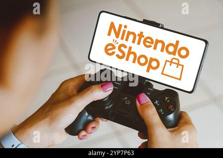 Dans cette illustration photo, le logo Nintendo eShop est affiché sur l'écran d'un smartphone lorsqu'une personne joue sur une manette de jeu Banque D'Images